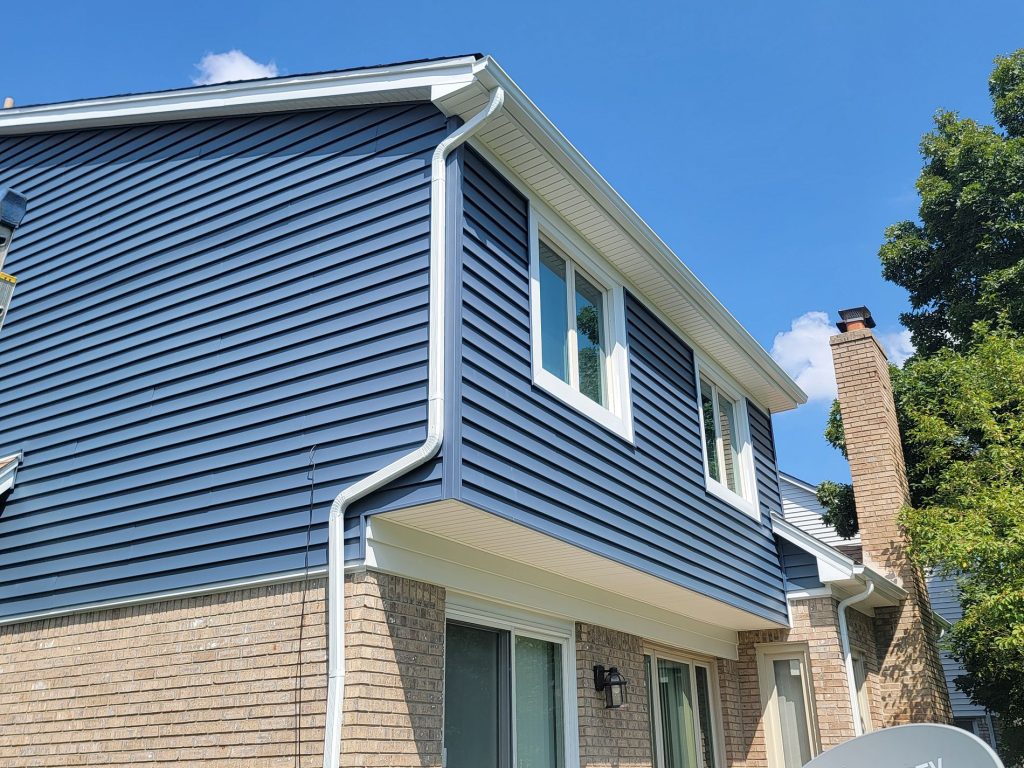 After: the same two-story home with deep blue Dutch lap siding, white window trim, white fascia and light vented soffit