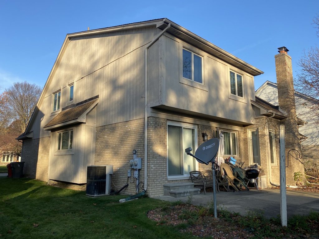 Before: rear of a two-story home with pale tan vertical board siding above tan brick and a patio