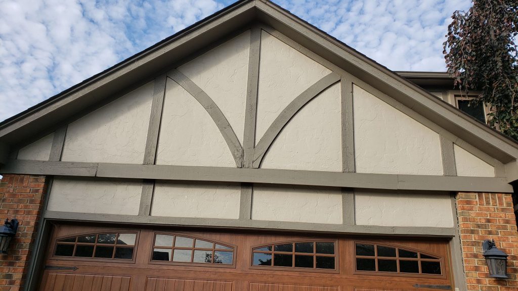 Before: Tudor-style garage gable with light textured panels and painted brown-gray trim boards above a wood-tone garage door