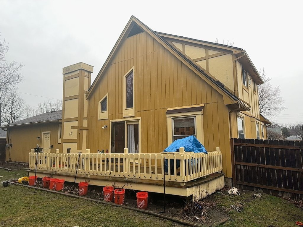 Before: rear of a Clinton Township home with mustard-yellow vertical board siding, a tall gable and a boxed chimney chase with Tudor-style trim